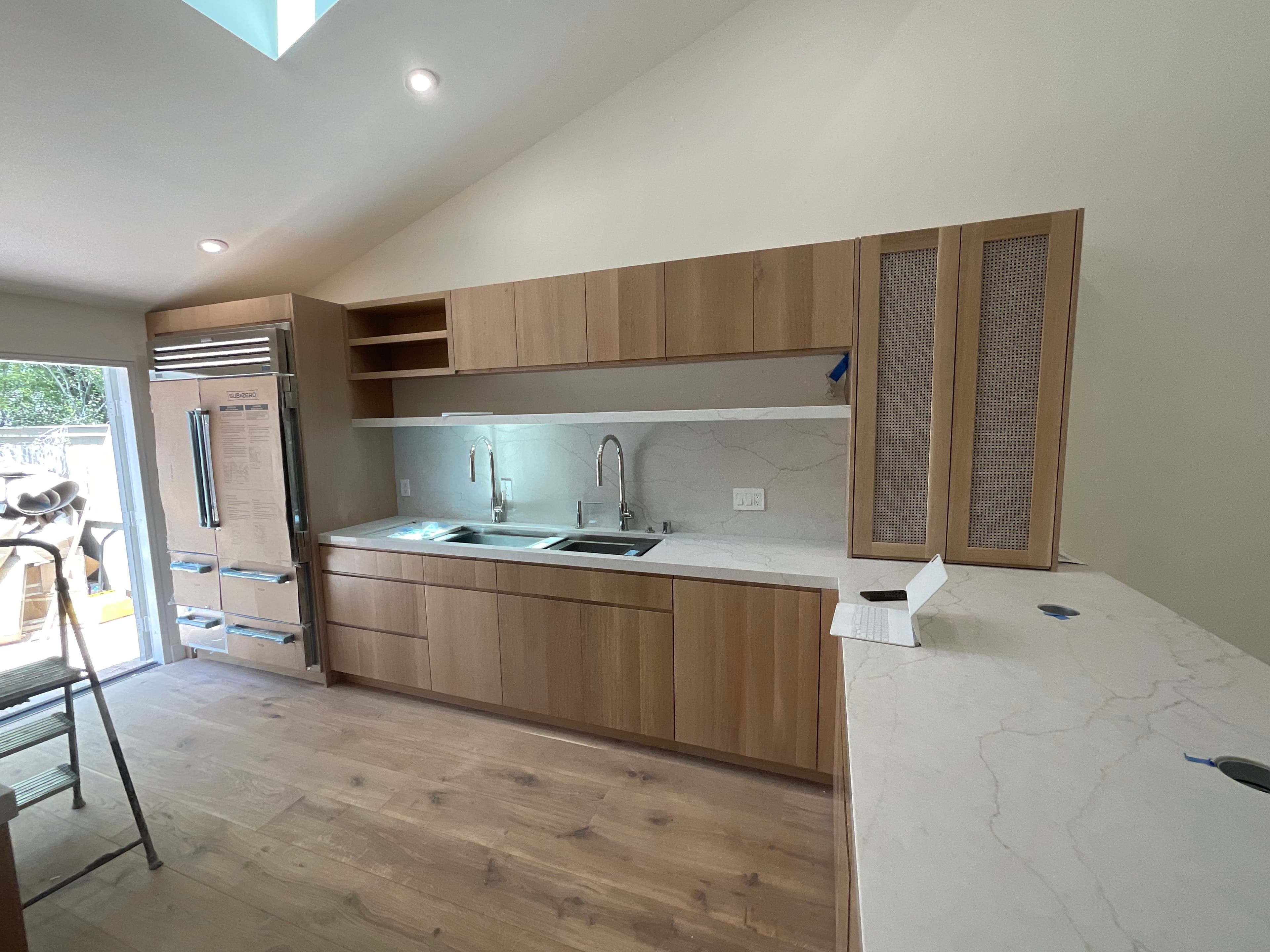 Modern kitchen featuring light wood cabinetry, white marble countertops, and double sinks under vaulted ceilings.
