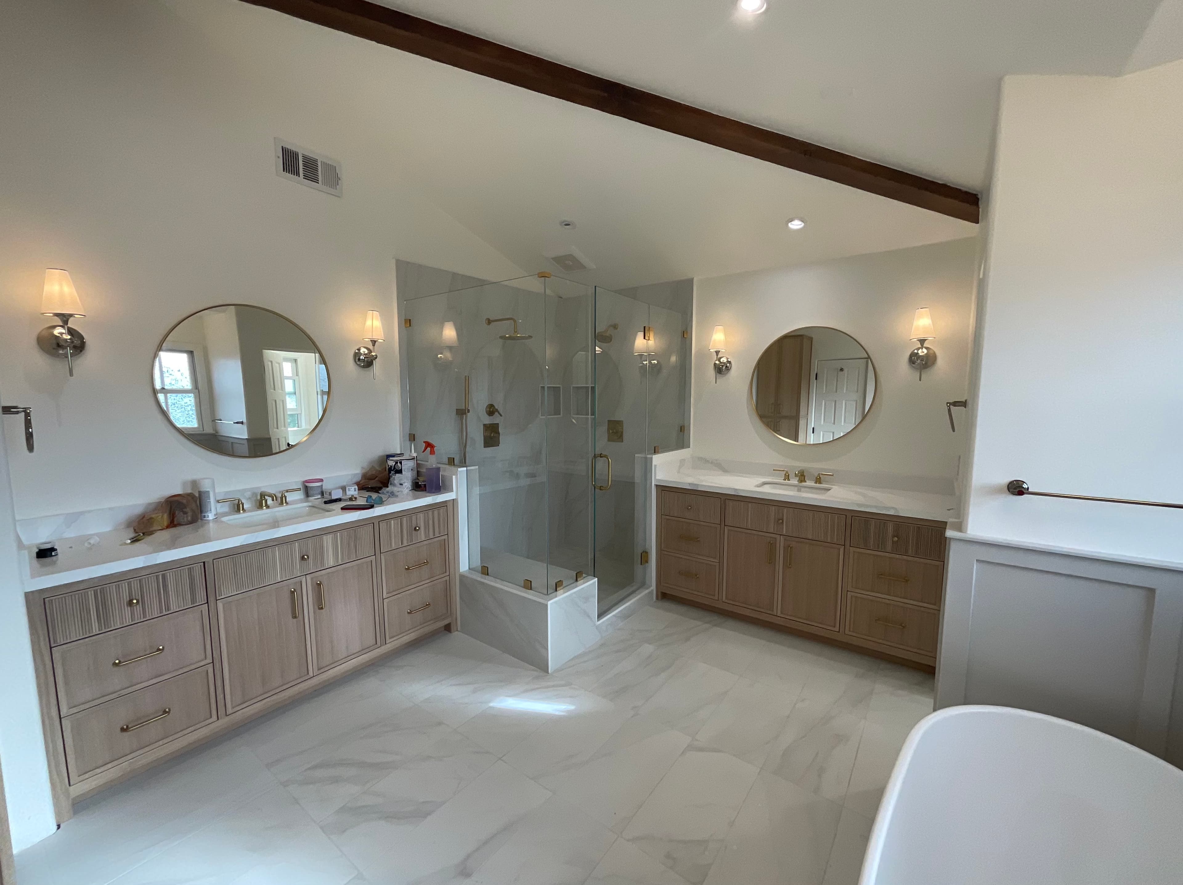 Modern bathroom with light wood vanities, round gold mirrors, and a glass-enclosed marble shower.
